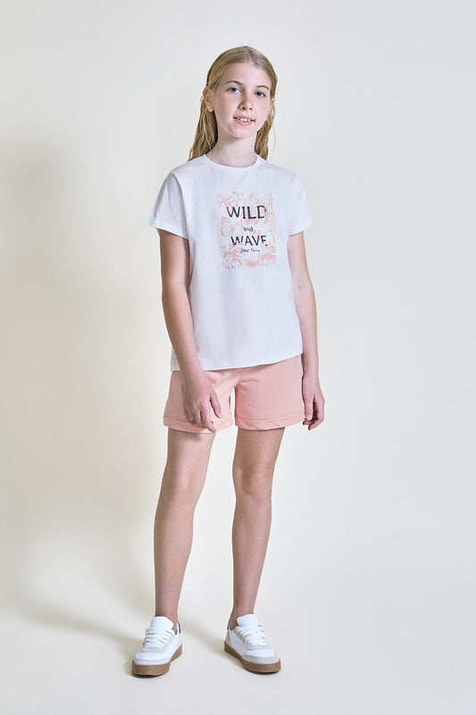 Niña con camiseta blanca de manga corta con estampado “Wild and Wave” en tonos suaves, combinada con short rosa, sobre fondo claro.