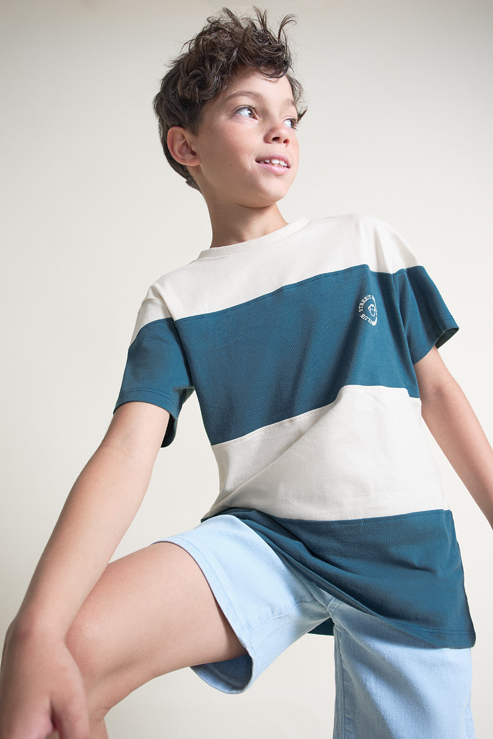 Niño con camiseta bicolor en tonos verde azulado y crudo de manga corta, combinada con short azul claro, sobre fondo neutro.
