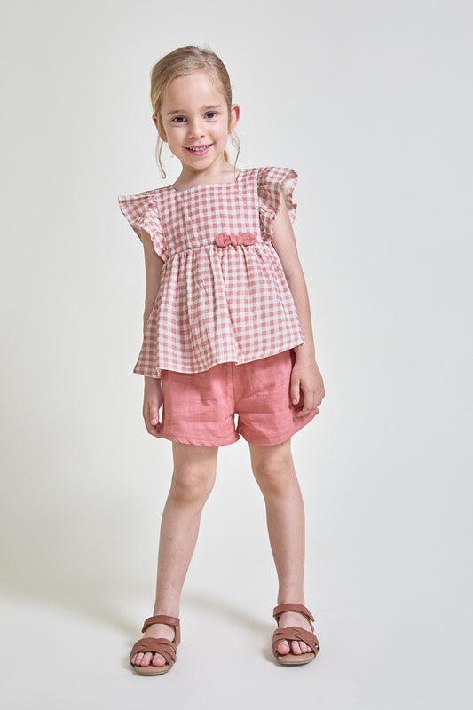Niña con conjunto de verano formado por blusa de cuadros vichy rosa con mangas de volante y detalle frontal, y short liso a juego, sobre fondo claro.