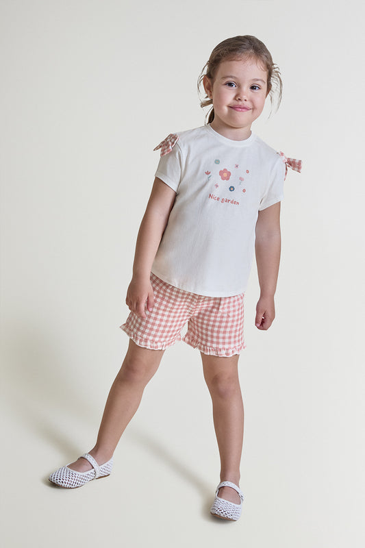 Niña con conjunto de verano formado por camiseta blanca con estampado floral y texto “Nice garden”, y short de cuadros vichy rosa con volante en el bajo, sobre fondo claro.