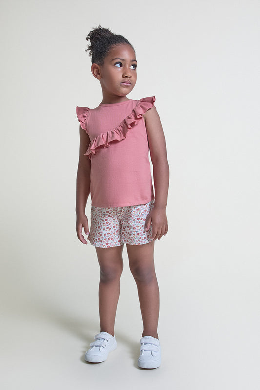 Niña con conjunto de verano formado por camiseta en tono teja con volante diagonal y short estampado floral multicolor, sobre fondo claro.