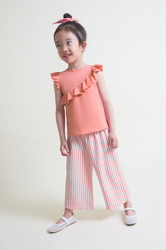 Niña con conjunto de verano compuesto por camiseta coral con volante diagonal y pantalón ancho de rayas en tonos coral y blanco, sobre fondo claro.