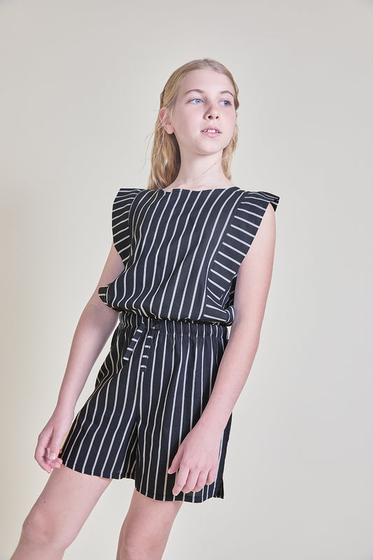 Niña con mono corto color marino de rayas blancas, sin mangas y con volante en los hombros, cintura elástica y lazada, ideal para verano.
