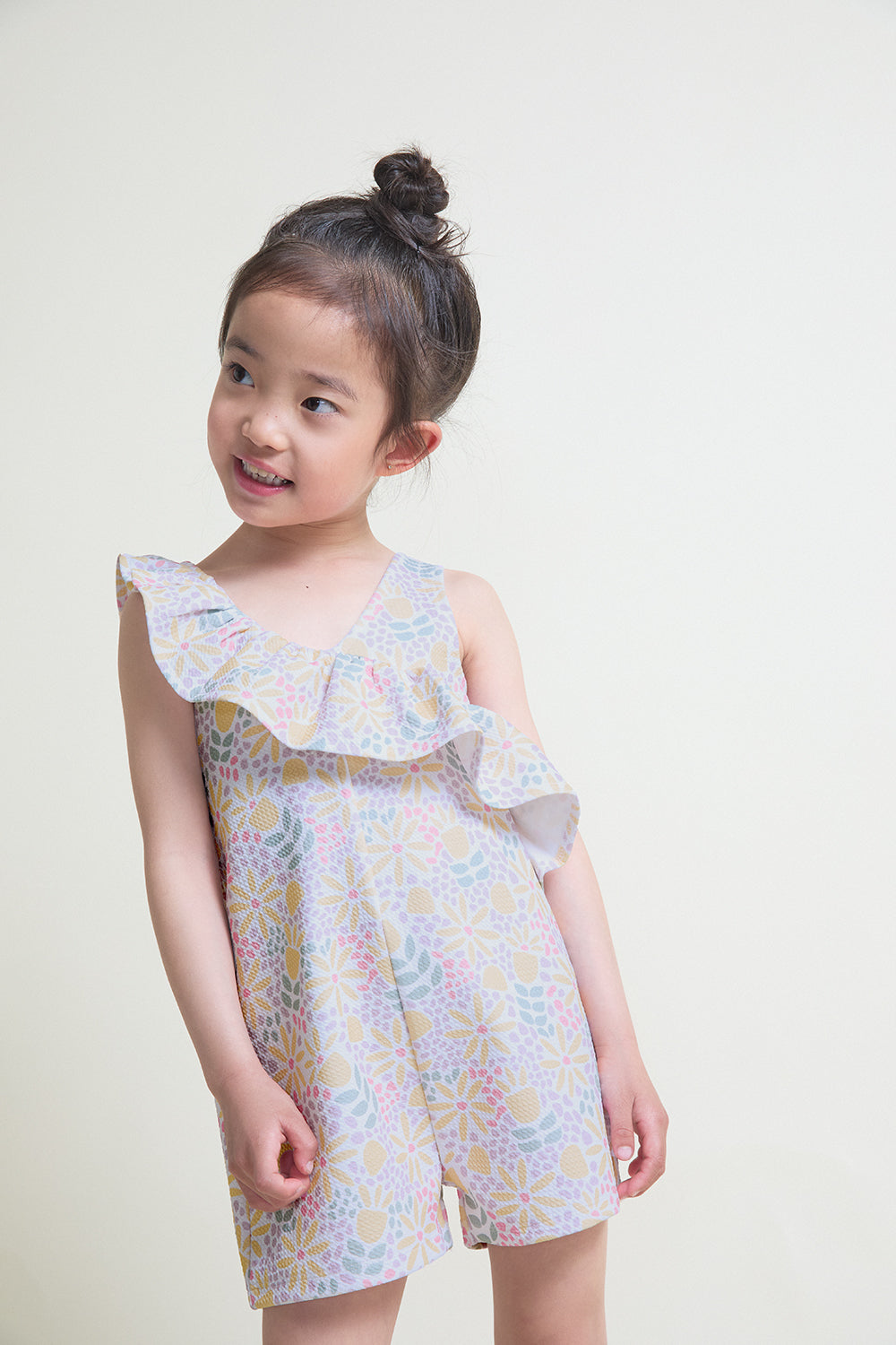Niña con mono corto de verano con volante asimétrico en el escote y estampado geométrico en tonos pastel sobre fondo claro.