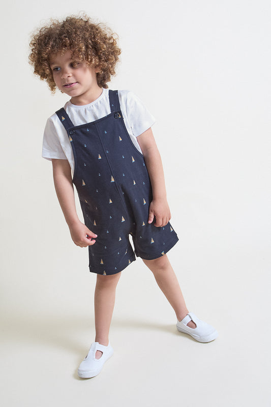 Niño con peto corto azul marino estampado con pequeños barquitos, combinado con camiseta blanca de manga corta, sobre fondo claro.