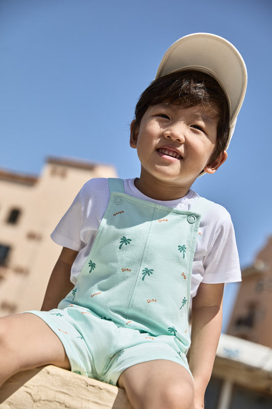 Niño con peto corto verde menta estampado con palmeras y detalles “Dino”, combinado con camiseta blanca, ideal para verano.