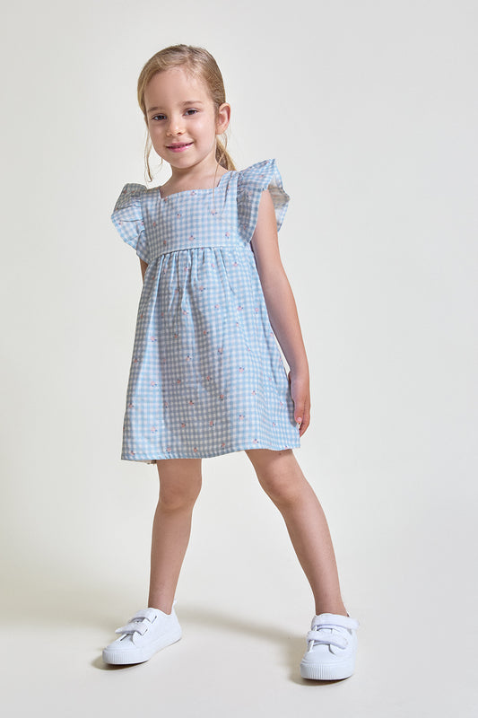 Niña con vestido de verano azul de cuadros vichy con pequeños motivos florales y mangas con volante, fotografiado sobre fondo claro.